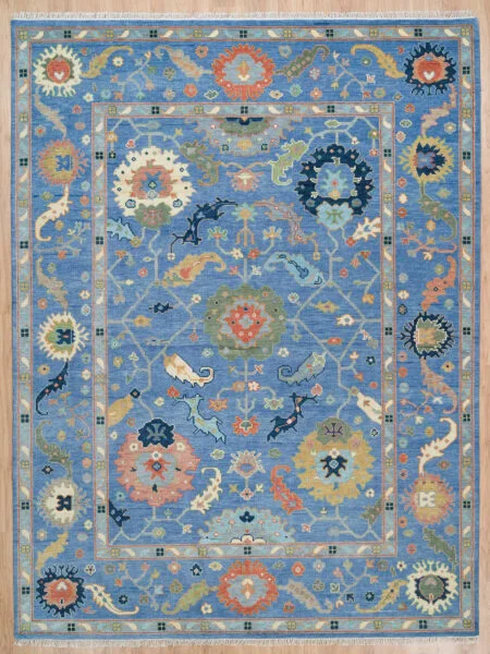 Hand-knotted wool Oushak rug with a blue field, wide blue border, and oversized rosette, palmette, vine, serrated-leaf, and cloud-band motifs in coral, gold, ivory, and sage.