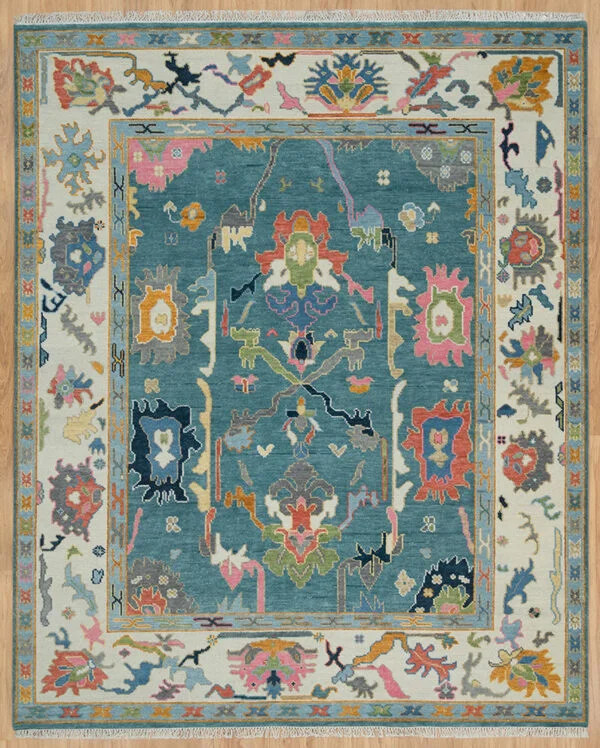 Hand-knotted wool Oushak rug with a teal field, wide ivory border, and oversized palmette, rosette, vine, and serrated-leaf motifs in blush, coral, navy, rust, olive, and gold.