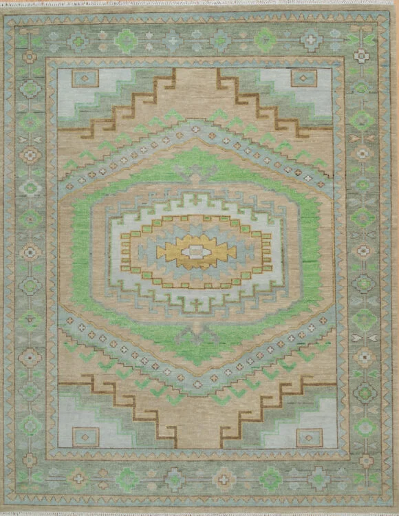 Hand-knotted wool oushak rug with a warm sand field, wide sage border, and a large oversized central stepped medallion with diamond and chevron motifs in green, blue, and gold.