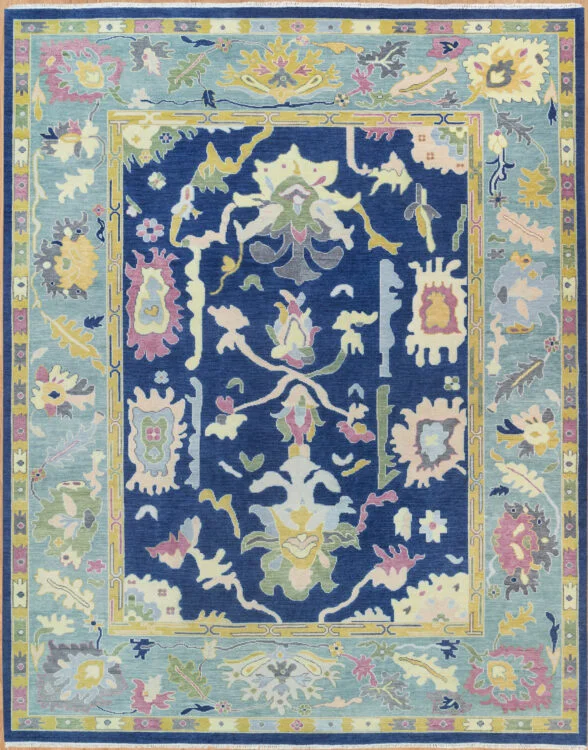 Hand-knotted wool oushak rug with a deep navy field, wide light blue border, and dense oversized palmettes and rosettes in pastel ivory, rose, and green with bold glowing contrast.