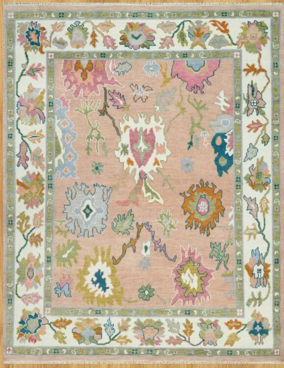 Hand-knotted wool Oushak rug with a peach field featuring oversized palmettes in pink, blue, olive, and coral, framed by a wide ivory vine border.