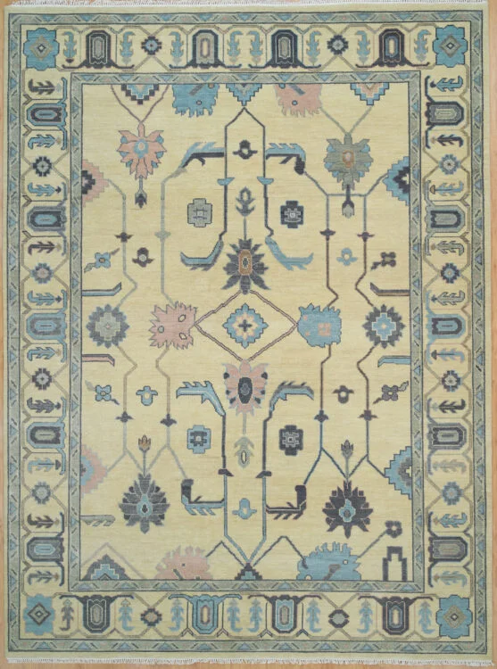 Hand-knotted wool Oushak rug with a sand field featuring angular vines and palmettes in charcoal and blue, framed by a wide pale blue geometric scroll border.