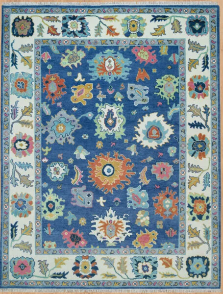 Hand-knotted wool Oushak rug with blue field, ivory border, and vivid large-scale floral palmettes in coral, green, gold, and pink.