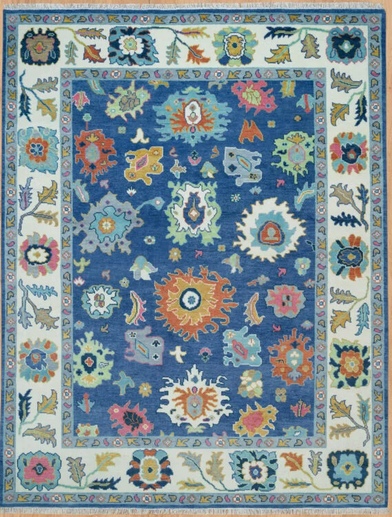 Hand-knotted wool Oushak rug with blue field, ivory border, and vivid large-scale floral palmettes in coral, green, gold, and pink.