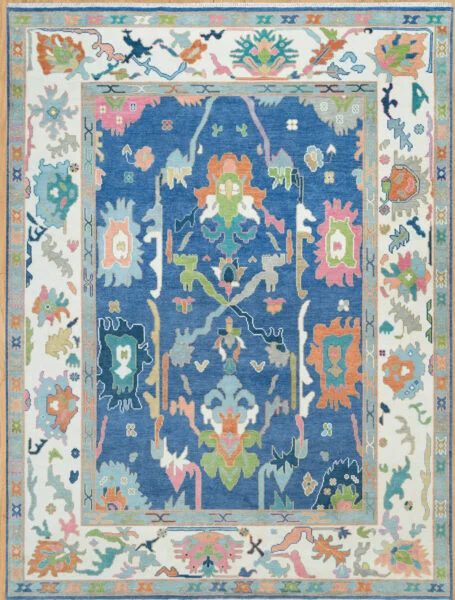 Hand-knotted wool Oushak rug with rich blue field, ivory border, and large floral palmettes in coral, green, peach, and ivory.