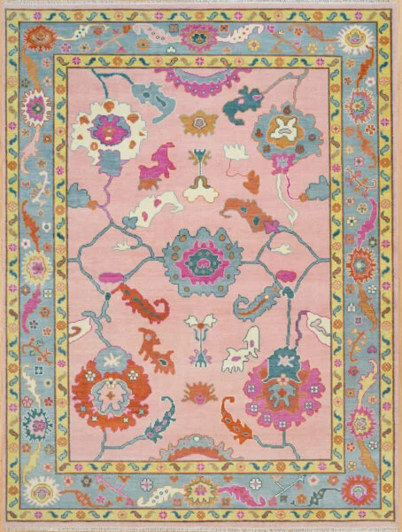 Hand-knotted wool Oushak rug with blush pink field, blue border, and vivid large-scale floral palmettes in rust, teal, gold, and blue.