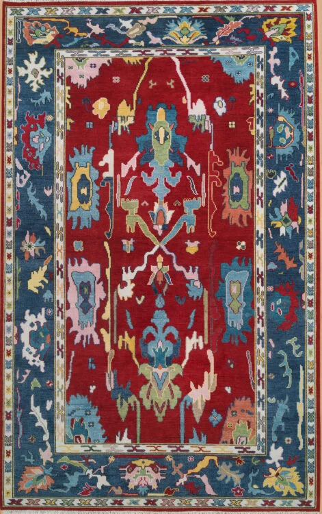 Hand-knotted modern Oushak wool rug with a rich red field, large central branching floral motif, and a wide navy border with colourful abstract palmettes.