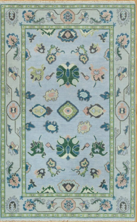 Hand-knotted wool oushak rug with a soft blue field, wide sage-green border, and balanced medium-scale palmettes and rosettes in green, blush, navy, and peach.