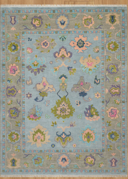 Hand-knotted wool oushak rug with a mid-blue field, wide taupe-green border, and balanced large-scale palmettes and rosettes in olive, blush, plum, and peach.