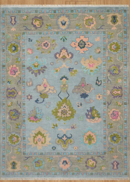 Hand-knotted wool oushak rug with a mid-blue field, wide taupe-green border, and balanced large-scale palmettes and rosettes in olive, blush, plum, and peach.